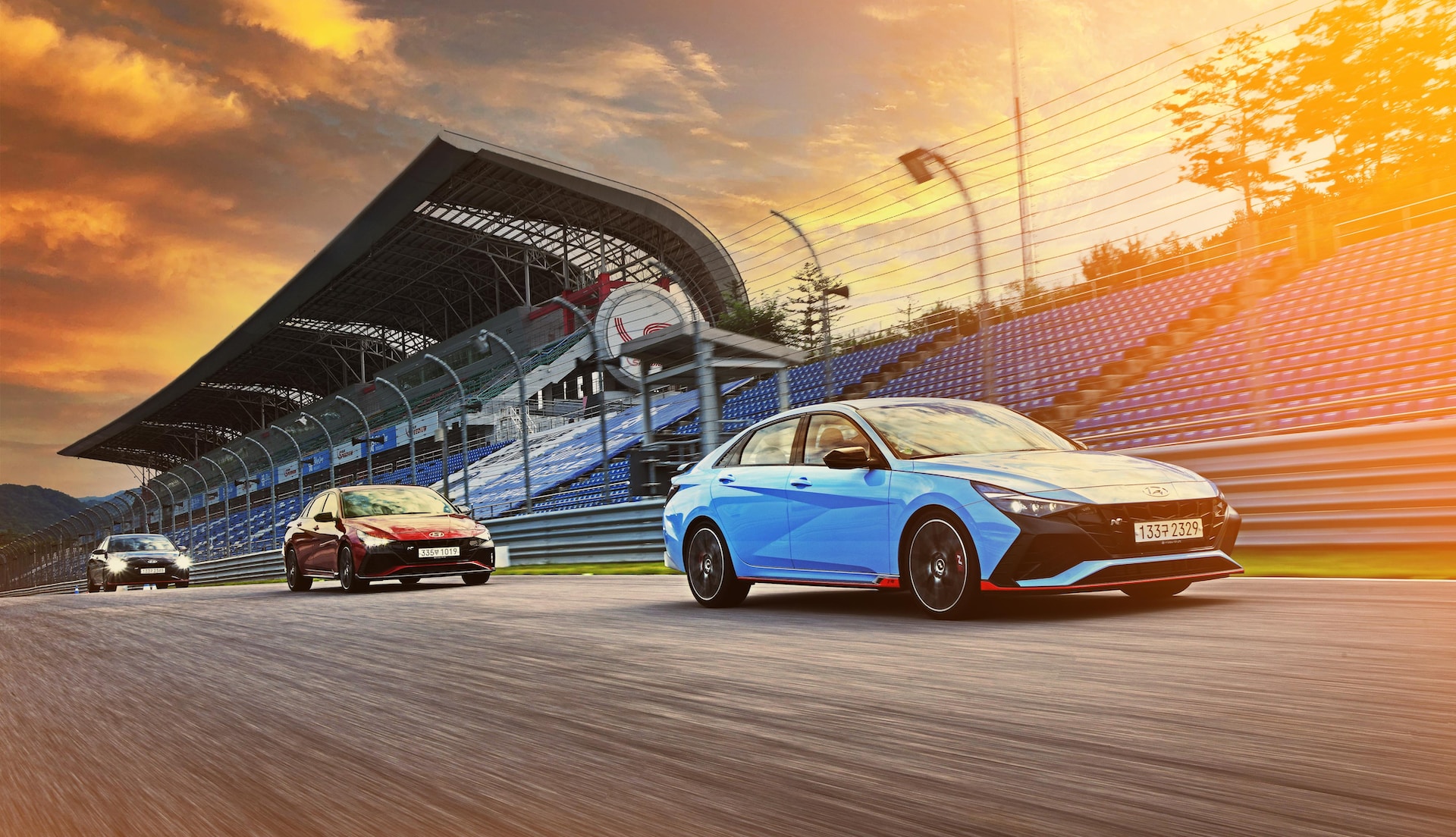 three Hyundai cars driving around a racetrack during sunset with the Blue one in the lead.